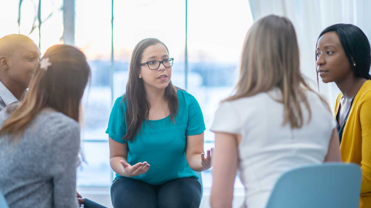A female Delegate with glasses and a green top hosts a meeting with four other people. She has a concerned expression on her face.