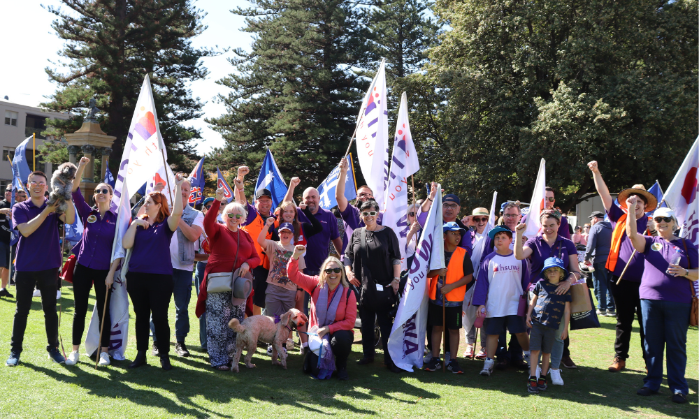 A group of HSUWA Members and Officials stand on the grass smiling at Fremantle Esplanade with their fists in the air. They hold flags and wear purple after the May Day March 2023.