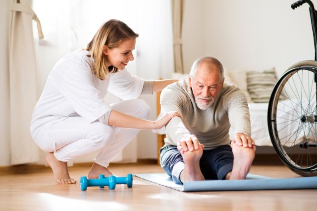 An aged care physiotherapists assists an elderly gentleman with a lower back stretch as he is seated on a mat.
