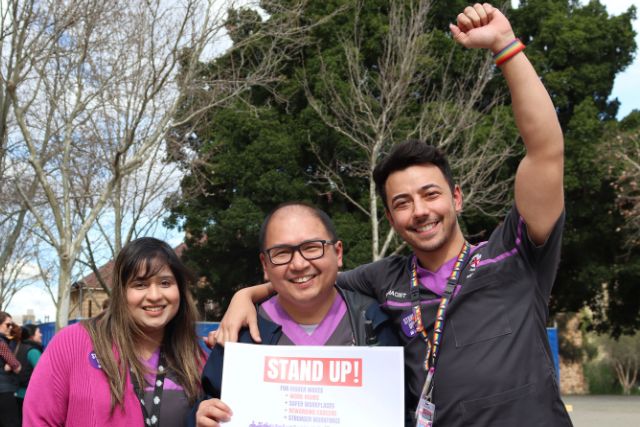 Three pharmacy members pose with a sign that says Stand Up!. The person on the right holds their fist in the air.