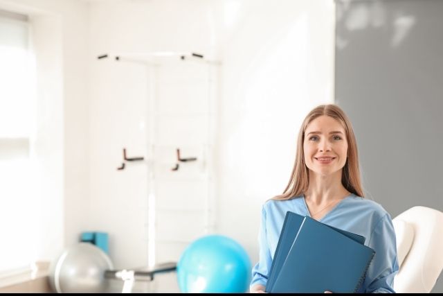 A blonde, female physiotherapist stands in front of equipment in her consult room.