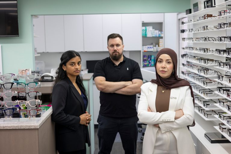 Three serious Optometrists stand with arms folded in front of racks of glasses.