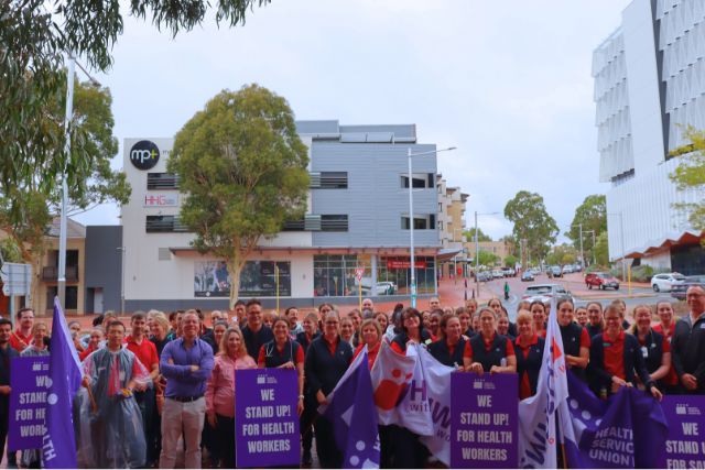 Allied Health Professionals stand at the front of Joondalup Health Campus with HSUWA signs and flags.