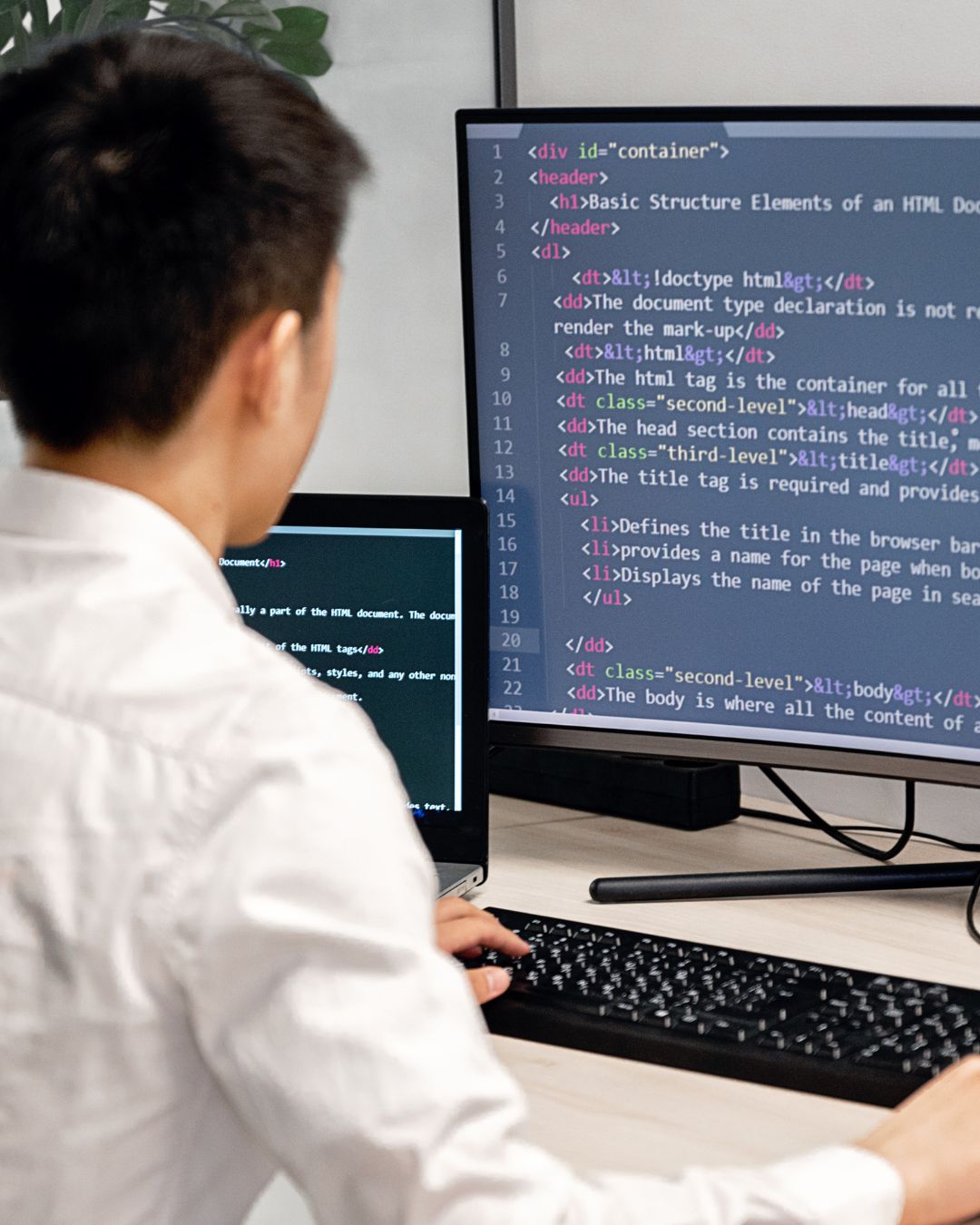 A person in a white shirt sits at a computer monitor with a curved screen with HTML code displayed.