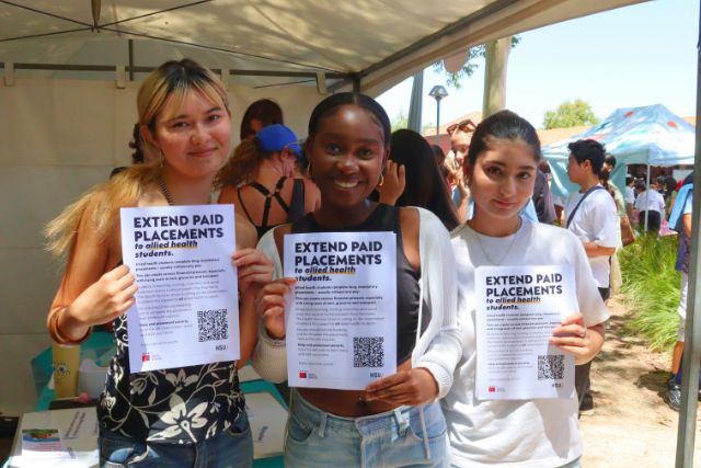 Three Allied Health Professional Students hold up the flyer to sign the Extend Paid Placements petition.