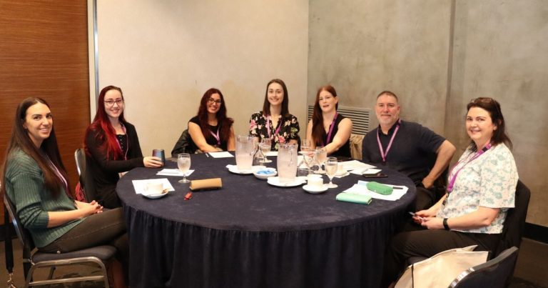 Seven HSUWA Delegates are seated at a table at the 2026 HSUWA Conference. They are smiling at the camera.