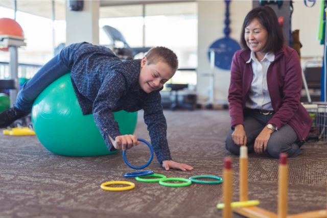 A boy with Down Syndrome reaches for coloured rings while supporting himself on a fit ball. His Occupational Therapists looks on smiling.